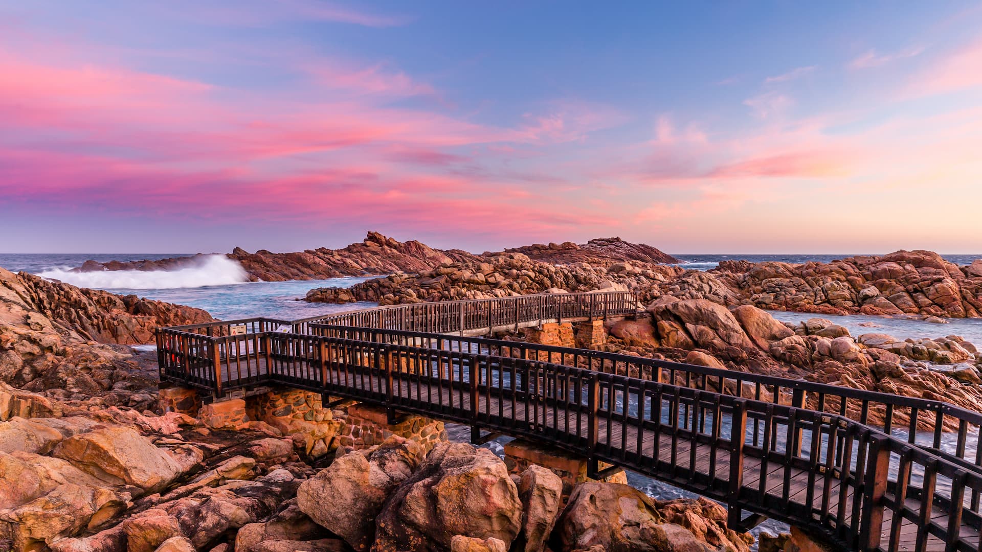 A wooden boardwalk extending over dark coastal rocks at sunset